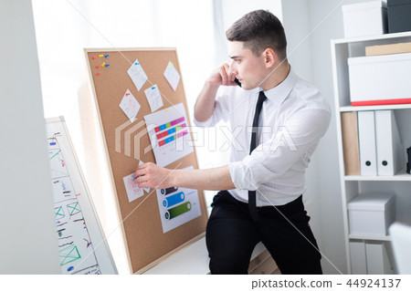 A young man in the office standing near the Board with stickers and talking on the phone. A young man in the office standing near the Board with stickers and talking on the phone. 44924137