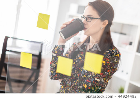 A young girl stands in the office near a transparent Board with stickers and drinks coffee. 44924482