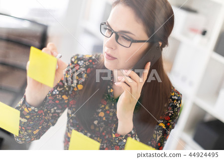 A young girl standing in the office near a transparent Board with stickers and talking on the phone. A young girl standing in the office near a transparent Board with stickers and talking on the phone. 44924495
