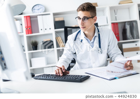 A young man in a white robe sitting at a table in the office. He holds a pen in his hand and works A young man in a white robe sitting at a table in the office. He holds a pen in his hand and works 44924830
