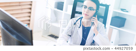 A beautiful young girl in a white robe is sitting at the table, talking on the phone and holding her 44927059