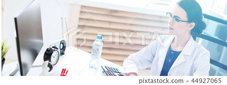 A beautiful young girl in a white robe is sitting at the table and typing on the keyboard. 44927065