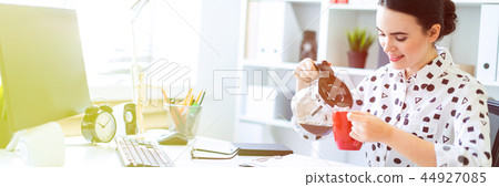 A young girl sits at a table in the office and pours coffee into a cup. 44927085