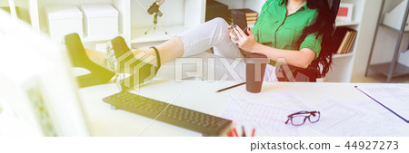 A young girl sits in the office, threw her legs on the table and holds the phone in her hands. 44927273