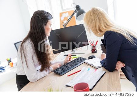 Two girls stand in the office near the table and look at the monitor. Two girls stand in the office near the table and look at the monitor. 44927580