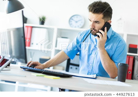 A young man standing in the office at the computer Desk and talking on the phone. A young man standing in the office at the computer Desk and talking on the phone. 44927613