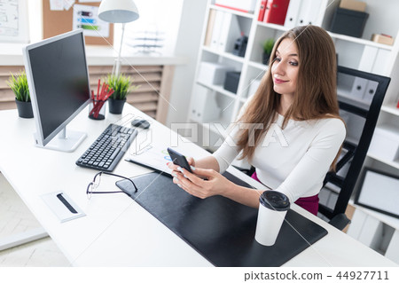 A young girl sitting at a table in the office and holding a phone. Next to the table is a white A young girl sitting at a table in the office and holding a phone. Next to the table is a white 44927711