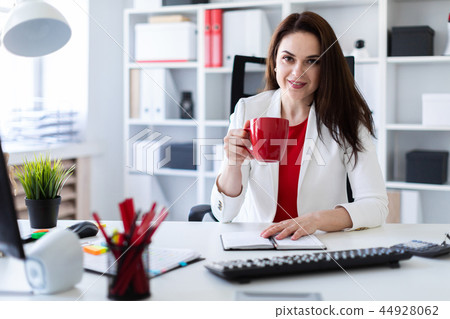 A young girl sitting in the office at the computer Desk and holding a red Cup. A young girl sitting in the office at the computer Desk and holding a red Cup. 44928062