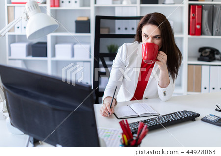 A young girl sitting in the office at the computer Desk and holding a red Cup. A young girl sitting in the office at the computer Desk and holding a red Cup. 44928063