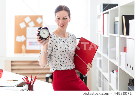 A young girl is standing in the office and holding a red folder with documents and an alarm clock. 44928071