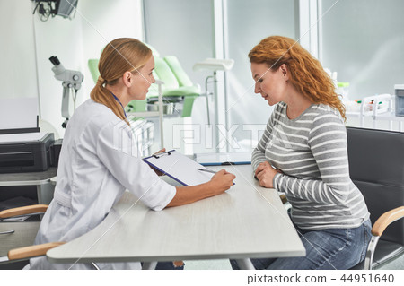 Red-haired lady looking at clipboard while sitting at the table with doctor Red-haired lady looking at clipboard while sitting at the table with doctor 44951640