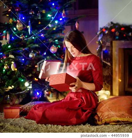 Beautiful young woman opening magical Christmas gift by a fireplace Beautiful young woman opening magical Christmas gift by a fireplace 44955939