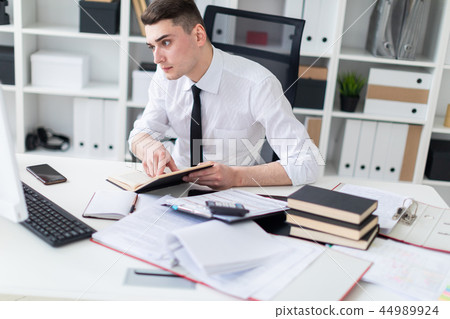 A young man working at a table in the office with a book, documents and a computer. 44989924