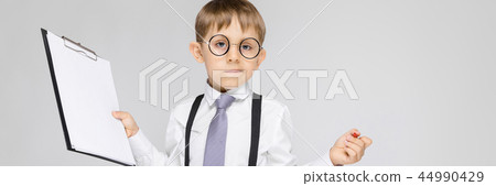 A charming boy in a white shirt, suspenders, a tie and light jeans stands on a gray background. The A charming boy in a white shirt, suspenders, a tie and light jeans stands on a gray background. The 44990429