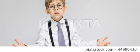A charming boy in a white shirt, suspenders, a tie and light jeans stands on a gray background. The A charming boy in a white shirt, suspenders, a tie and light jeans stands on a gray background. The 44990430