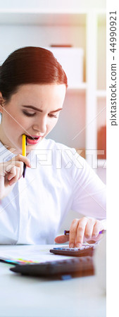 A young girl in the office holds a pen in her mouth and works with a calculator, documents and a 44990921