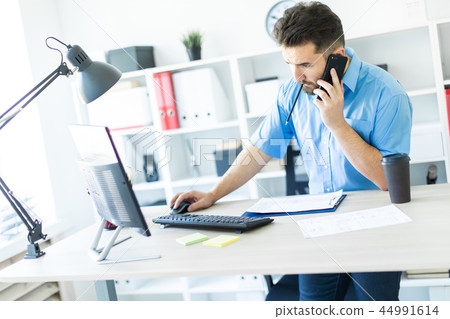 A young man standing in the office at the computer Desk and talking on the phone. 44991614