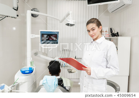 Charming beautiful girl dentist looks into the camera. Young African American male patient at chair 45009654