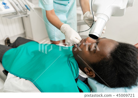 Woman dentist using x ray machine, patient lying on chair in dentistry. Young African American male 45009721