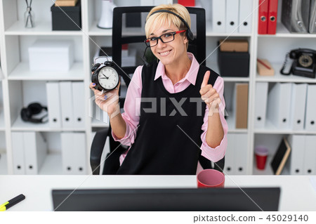 A young girl sitting at a computer table and holding an alarm clock. 45029140