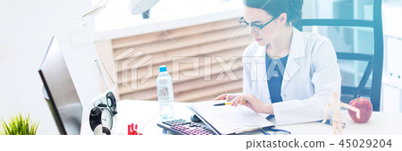 A beautiful young girl in a white robe is sitting at a computer desk with documents and a pen in her 45029204