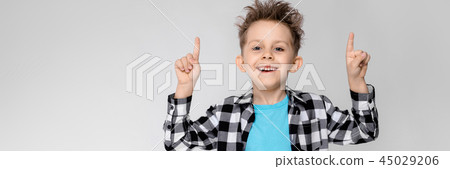 A handsome boy in a plaid shirt, blue shirt and jeans stands on a gray background. Boy smiling and A handsome boy in a plaid shirt, blue shirt and jeans stands on a gray background. Boy smiling and 45029206