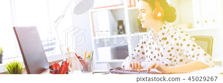 Beautiful young girl sitting in headphones at desk in office and typing on keyboard. 45029267