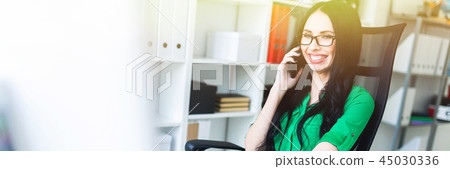 Smiling Young girl with glasses in the office speaks on the phone and holds a hand on the keyboard. 45030336