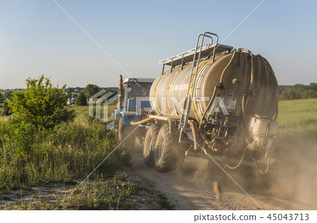 A blue tractor with a tank rides along a road in a wheat field. A blue tractor with a tank rides along a road in a wheat field. 45043713
