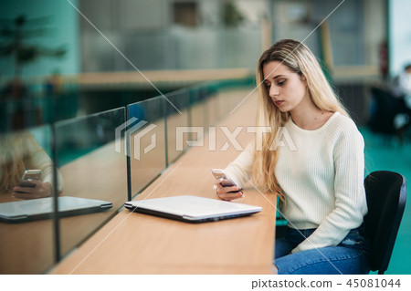 Blonde girl use phone in library. sudent sist on stool by the window 45081044