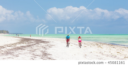 Active sporty tourist couple cycling down picture perfect white sand tropical beach of Paje Active sporty tourist couple cycling down picture perfect white sand tropical beach of Paje 45081161