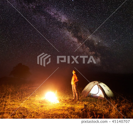 Young female tourist standing near illuminated tent, camping in mountains at night under starry sky Young female tourist standing near illuminated tent, camping in mountains at night under starry sky 45140707