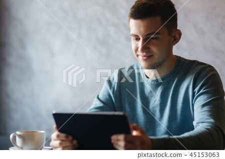 Guy is sitting in a cafe and using a tablet. Student spend time in cafe and drink coffee. He study 45153063