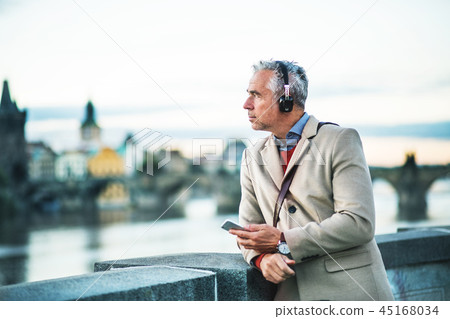 Mature businessman with headphones and smartphone standing by river in city. Mature businessman with headphones and smartphone standing by river in city. 45168034