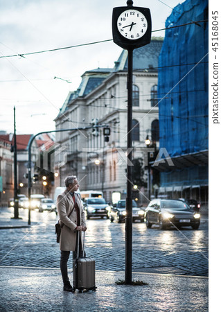 Mature businessman with suitcase standing by a road in city early in the morning. 45168045