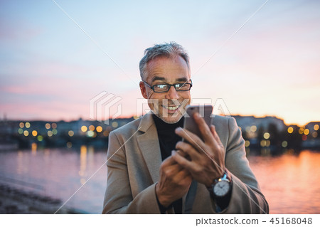 Mature businessman with smartphone standing by river in Prague city, taking selfie. 45168048
