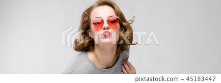 Charming young girl in a gray T-shirt on a gray background. Happy girl in red glasses Charming young girl in a gray T-shirt on a gray background. Happy girl in red glasses 45183447