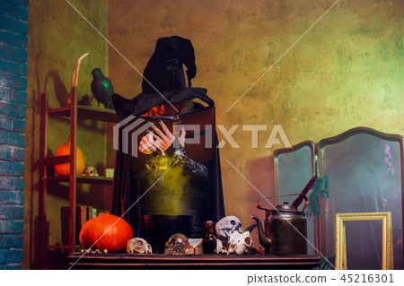 Photo of smiling witch in black hat reading spell over pot of green steam Photo of smiling witch in black hat reading spell over pot of green steam 45216301