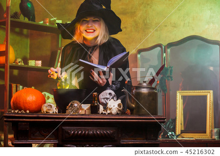 Portrait of smiling witch in black hat reading spell over pot with green steam 45216302
