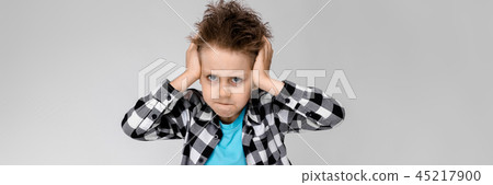 A handsome boy in a plaid shirt, blue shirt and jeans stands on a gray background. The boy folded A handsome boy in a plaid shirt, blue shirt and jeans stands on a gray background. The boy folded 45217900