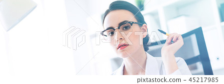 A beautiful young girl in a white robe is sitting at a computer desk with documents and a pen in her 45217985