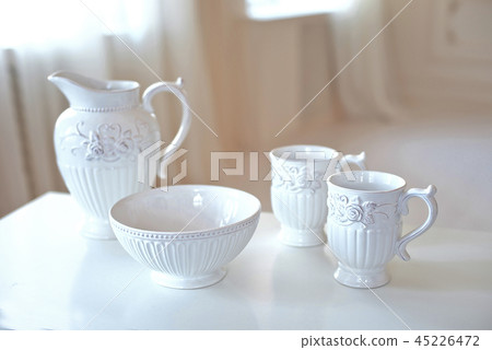Set of white ceramic kitchen utensils, dishes on white table in big bright room infront of window. Set of white ceramic kitchen utensils, dishes on white table in big bright room infront of window. 45226472