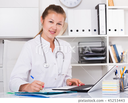 Young doctor woman in uniform is working behind laptop with documents Young doctor woman in uniform is working behind laptop with documents 45253443