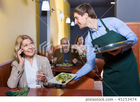 Positive waiter bringing order to charming smiling mature female Positive waiter bringing order to charming smiling mature female 45253474