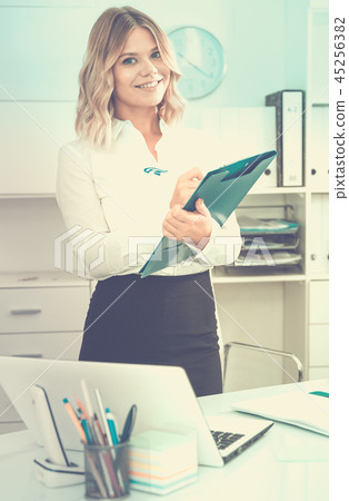 Well-wishing girl with folder of documents is standing in well-lit office Well-wishing girl with folder of documents is standing in well-lit office 45256382