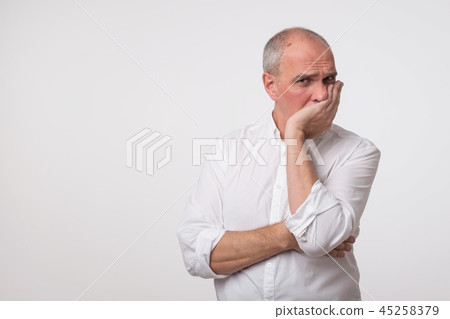 Displeased mature man in white shirt covering his face with hand over gray background. Displeased mature man in white shirt covering his face with hand over gray background. 45258379