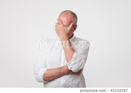 Displeased mature man in white shirt covering his face with hand over gray background. Displeased mature man in white shirt covering his face with hand over gray background. 45258387