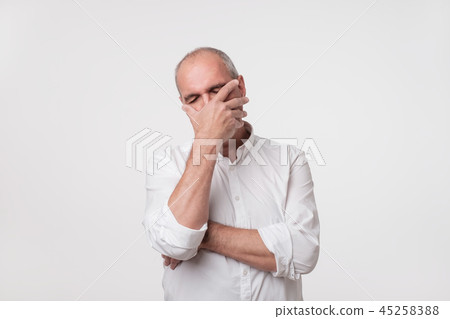 Displeased mature man in white shirt covering his face with hand over gray background. Displeased mature man in white shirt covering his face with hand over gray background. 45258388