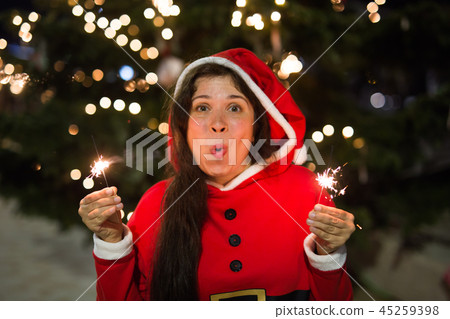 Holiday, Christmas and people concept - Young happy woman wearing Santa suit holding bengal light 45259398