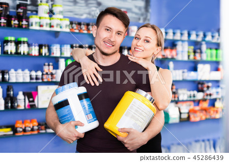Loving athletic couple holding plastic jars of sport food supplements in shop interior 45286439